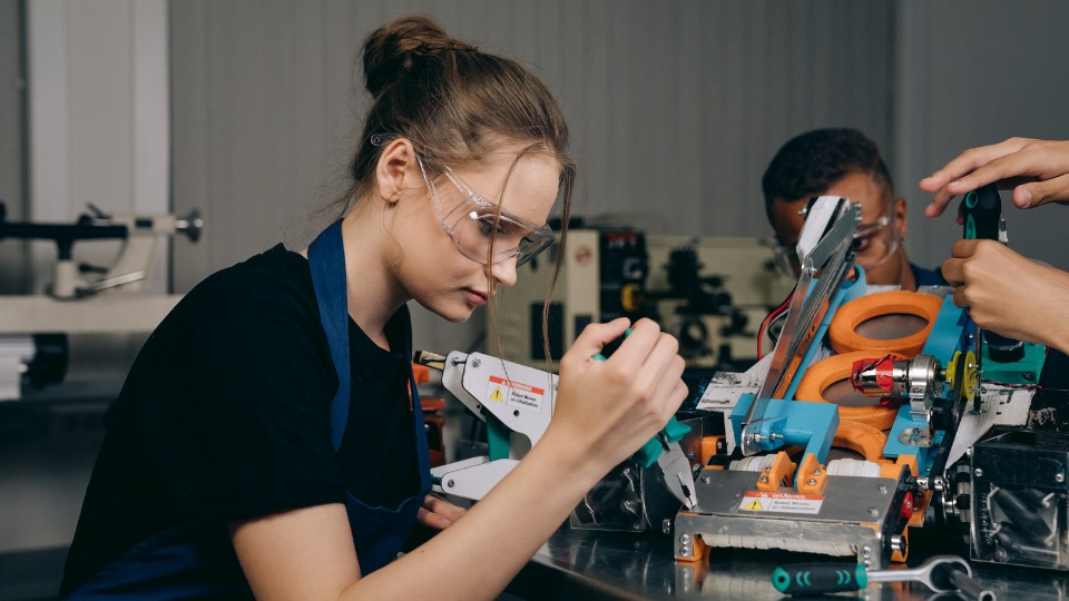 Young woman in safety goggles working on a mechanical project as part of a hands-on apprenticeship or training course, with other students in the background
