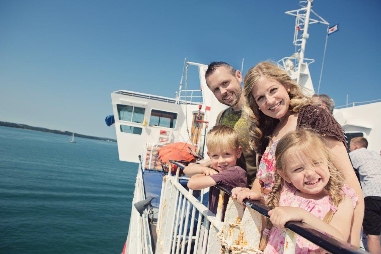 A family - two adults, two children - look over the side of a ferry to the camera. The ferry is at sea.