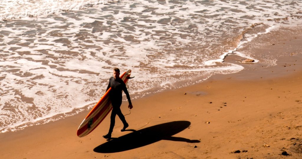 Man in a wetsuit carrying a surfboard along the shoreline at Compton Bay, Isle of Wight, with waves rolling in behind him