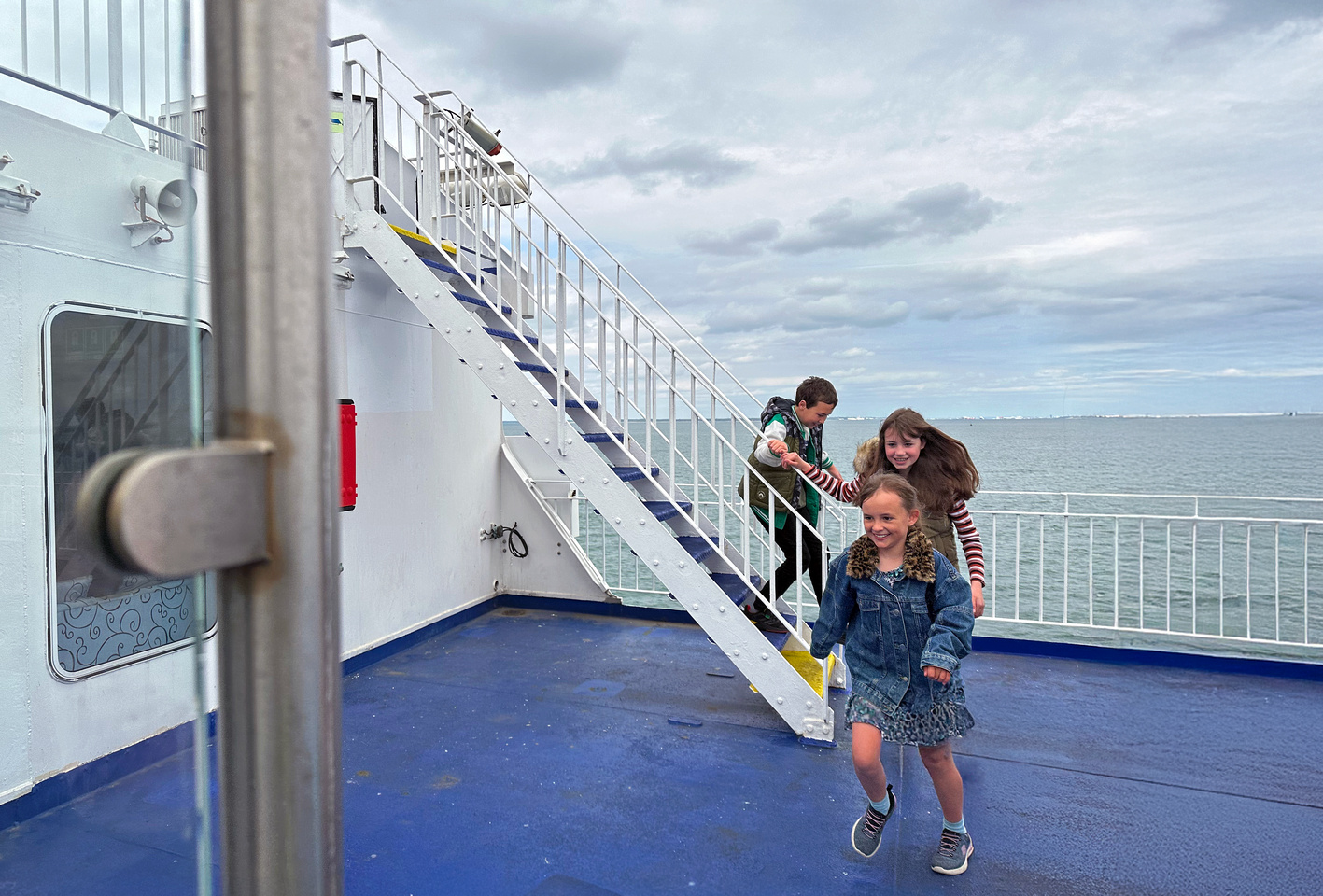 Three smiling children playing on the outside deck of a Wightlink ferry, with the sea and cloudy sky in the background.
