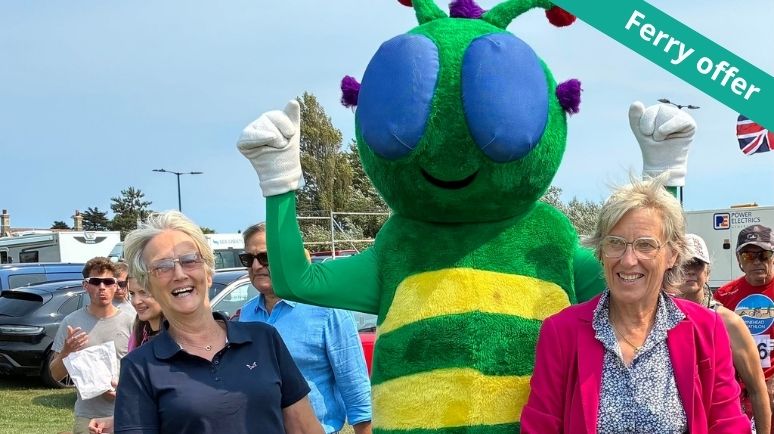 Green insect mascot posing with two smiling visitors at an outdoor event, with a crowd gathered behind them and a "Ferry offer" banner in the top corner