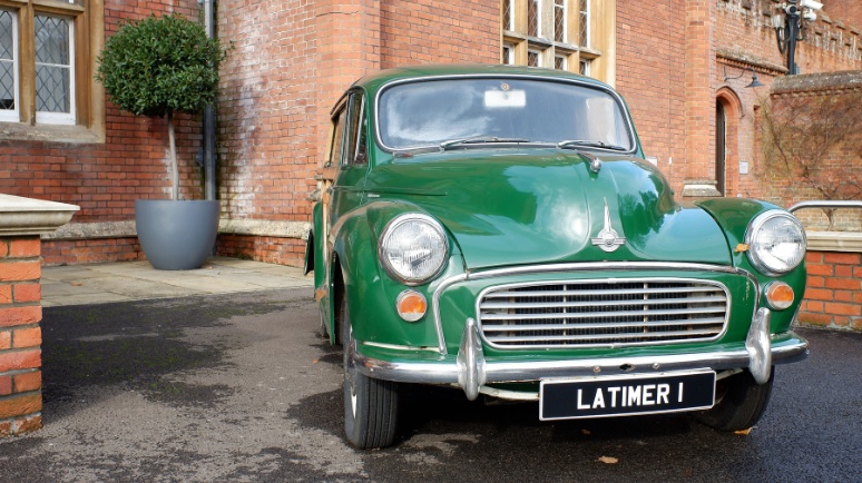 A classic green Morris Minor parked in front of a red-brick building with tall windows and a potted topiary tree nearby.