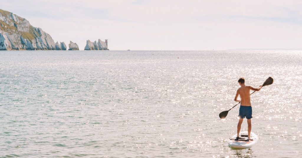 Man paddleboarding on calm water near the Needles on the Isle of Wight, with the iconic chalk stacks and lighthouse visible in the background.