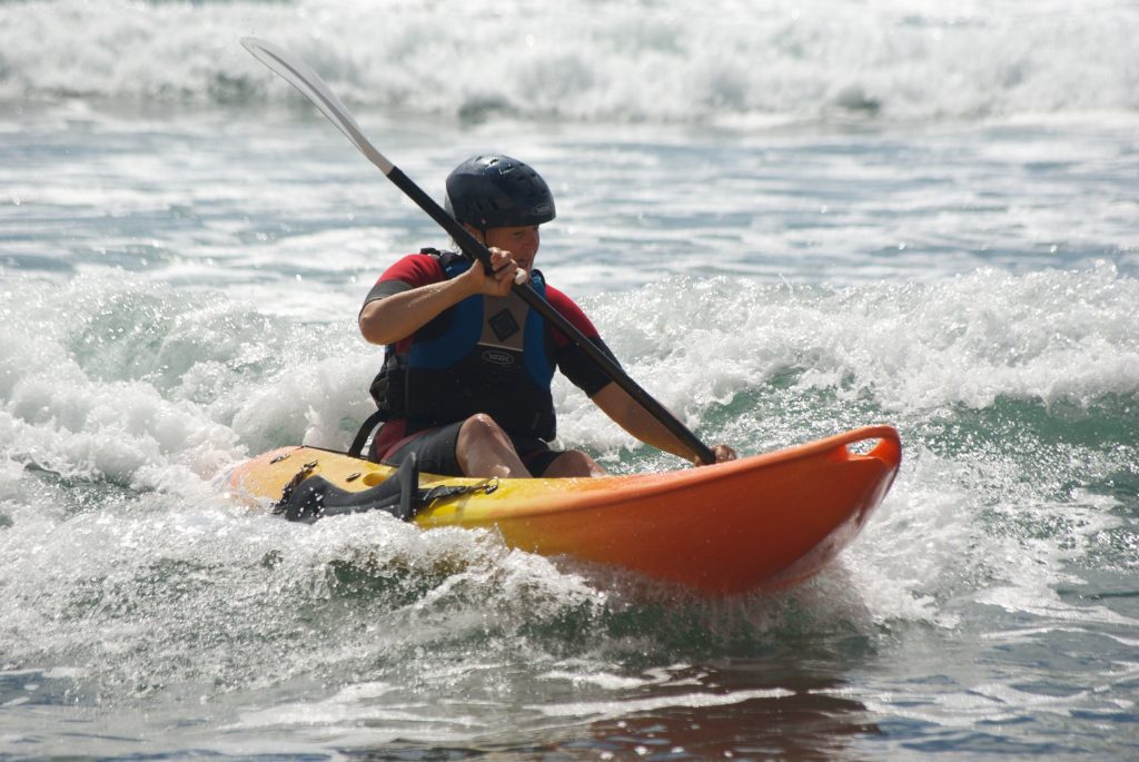 A person wearing a helmet and life jacket paddles a bright orange kayak through small breaking waves off the shores of the Isle of Wight, with white water splashing around them.