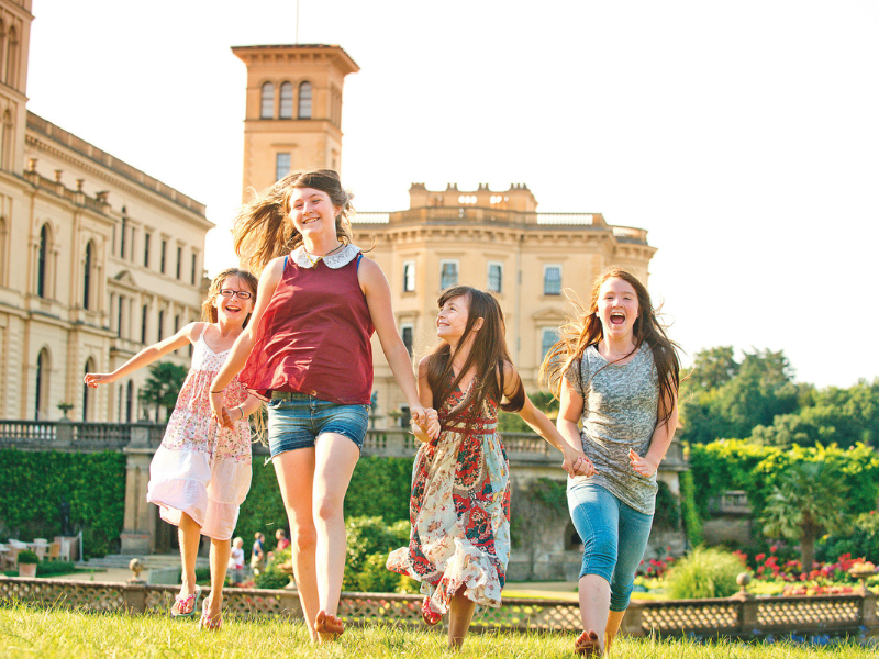 A group of children run through the gardens of Osborne on the Isle of Wight. The sun is shining and Osborne house is in the background