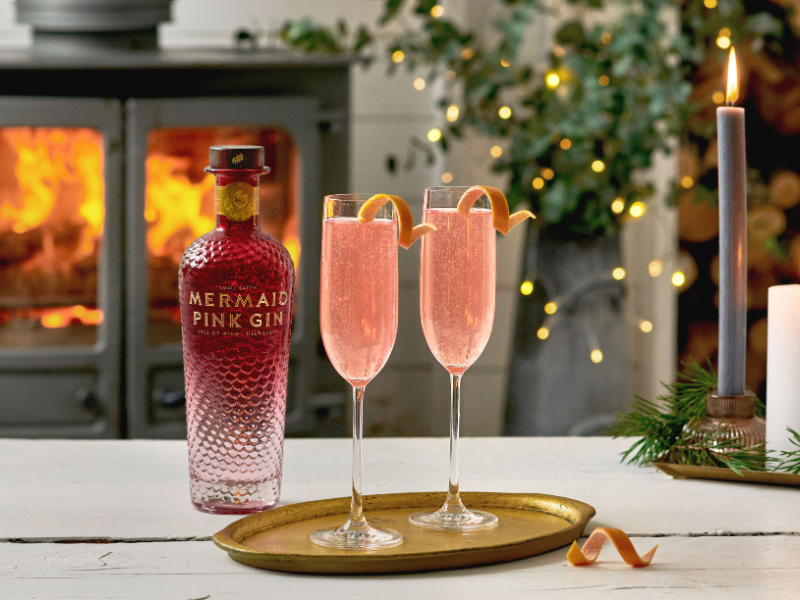 Two champagne flutes holding a cocktail and a bottle of Mermaid Pink Gin, against a festive backdrop of fairy lights, branches and a stove