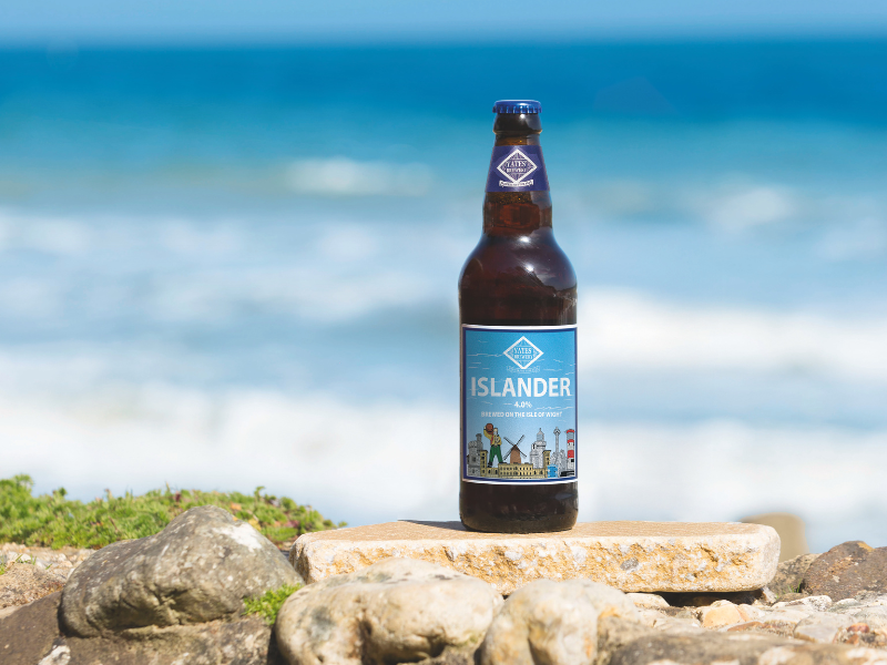 A brown bottle of craft ale, with a blue label that reads 'Islander' is perched on a stone in front of a beach