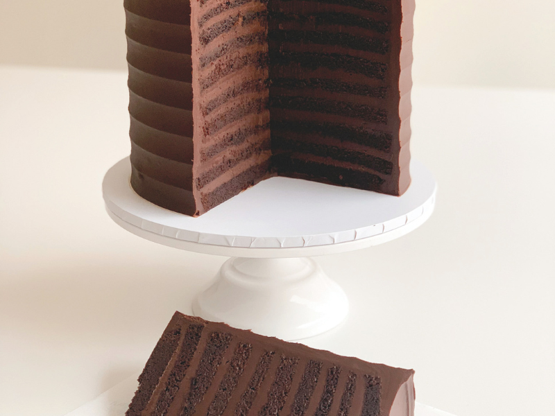 A white cake stand holds a multi-layer chocolate cake. There's a slice cut out of it and it's in the foreground on a plate