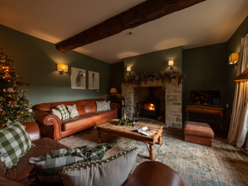 A cosy beamed living room with rich warm colours (green and tan), a fireplace, holly decorations and a Christmas tree