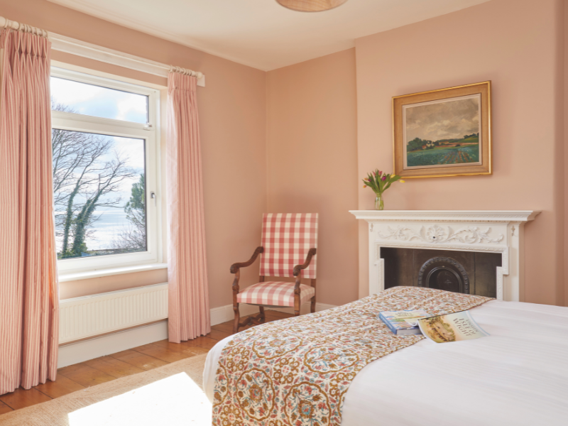 A pretty pink bedroom overlooking the sea, there's a bed, a fireplace, a chair and a picture on the wall