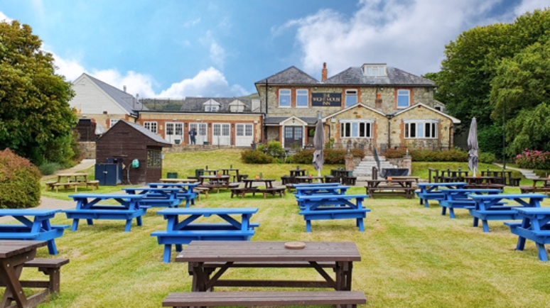 The Wight Mouse Inn with its large garden and rows of blue and brown picnic benches set out on the lawn, surrounded by trees under a partly cloudy sky.
