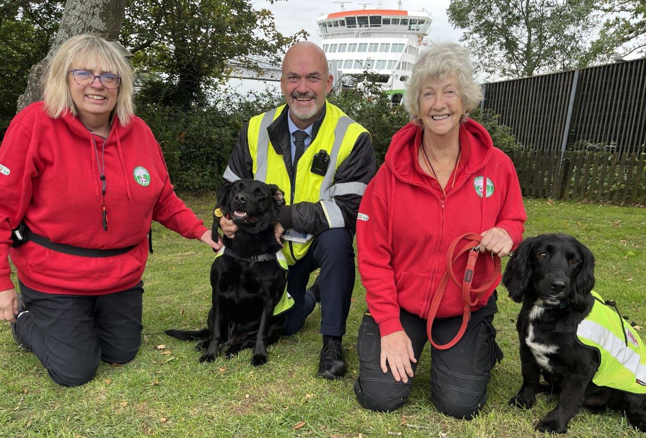 Three people - two from Wight SAR and one from Wightlink - pose with rescue dogs in front of a Wightlink ferry