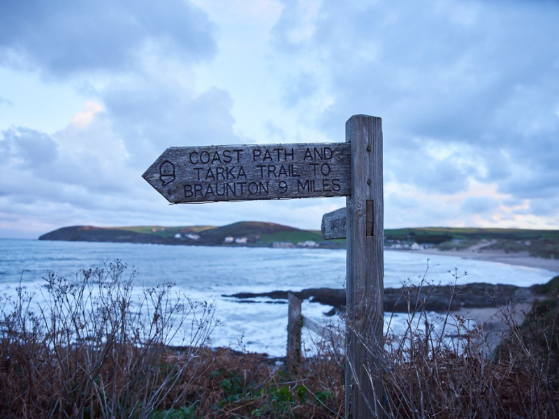 A wooden signpost, with pointed sign for a coastal path, against a dramatic coastal backdrop - image thanks to VisitBritain
