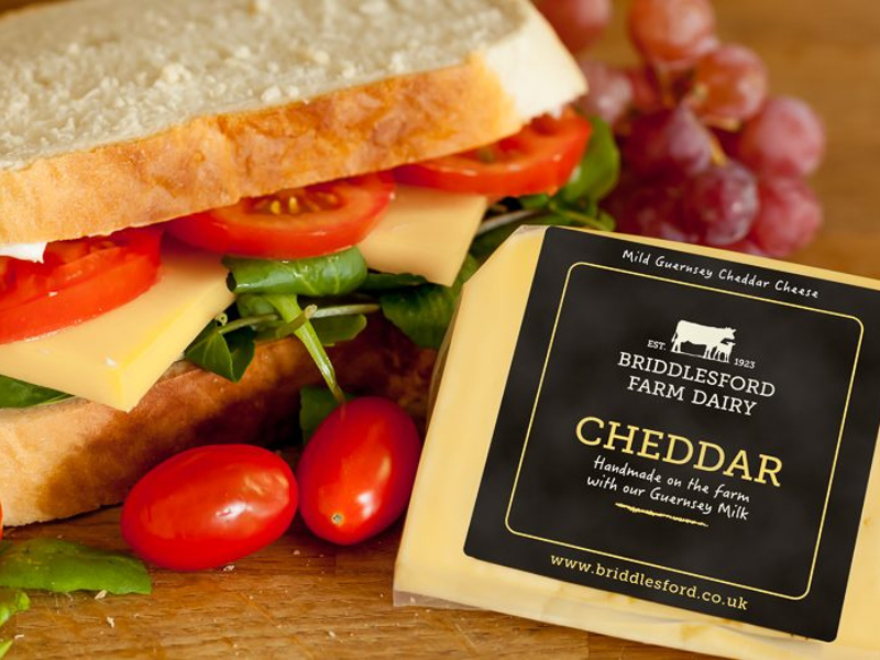 A cheese, tomato and rocket sandwich on white bread is on a chopping board alongside a packet of cheddar cheese