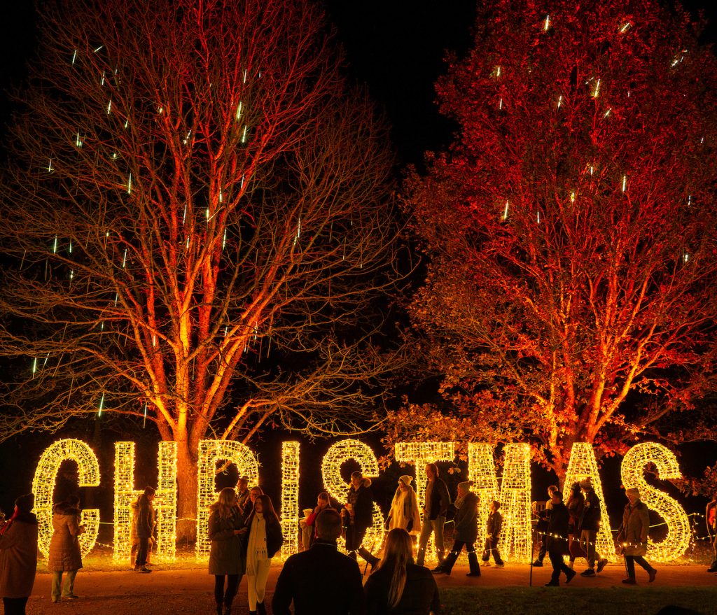 The word 'Christmas' in large lit-up letters stands beneath trees at a garden. It's night and there are people walking in front