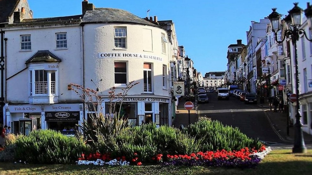 Street view in Ryde town centre with a white café building, fish and chip shop frontage, floral roundabout and cars parked along the high street.