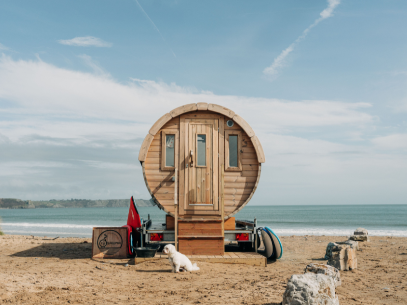 A round wooden mobile sauna on a sandy beach, there's a dog in front