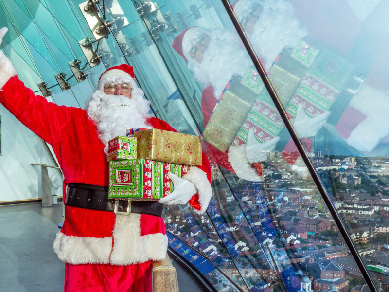 Father Christmas holding a pile of presents and waving, inside the Spinnaker Tower against the viewing window