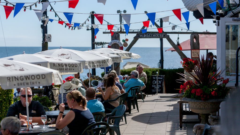 Outdoor seating area at a seaside pub with customers dining under Ringwood Brewery parasols, colourful bunting overhead and the sea in the background at The Spyglass Inn, Ventnor.