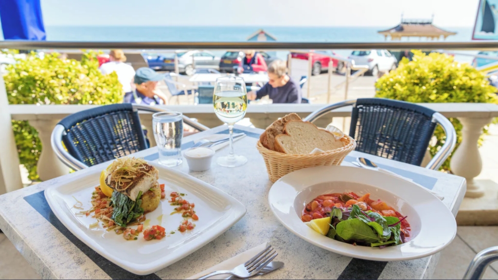 A table set with fresh seafood dishes, bread and a glass of wine on a sunny outdoor terrace overlooking the sea at a coastal restaurant on the Isle of Wight.