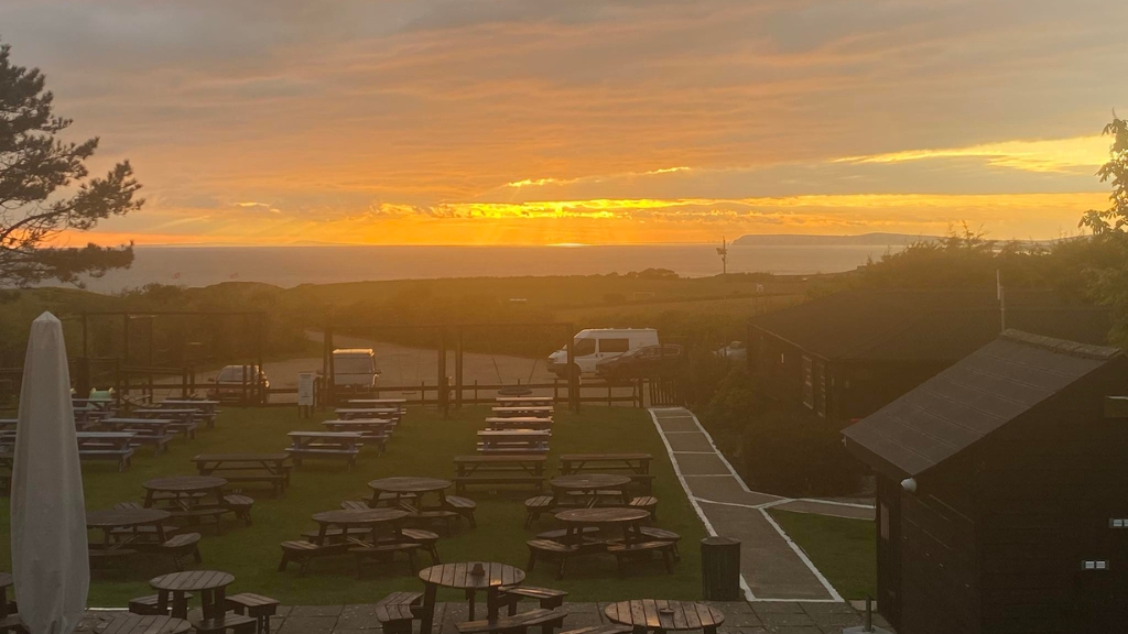 Large pub garden at The Wight Mouse Inn in Chale with picnic benches overlooking a dramatic sunset across rolling countryside and the sea.