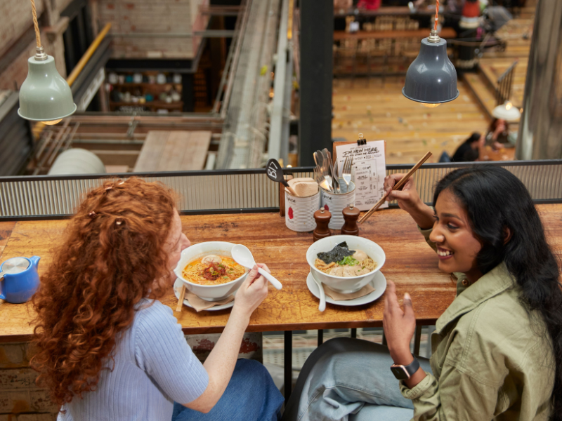Two friends eating bowls of food on a mezzanine of a restaurant - Credit VisitBritain