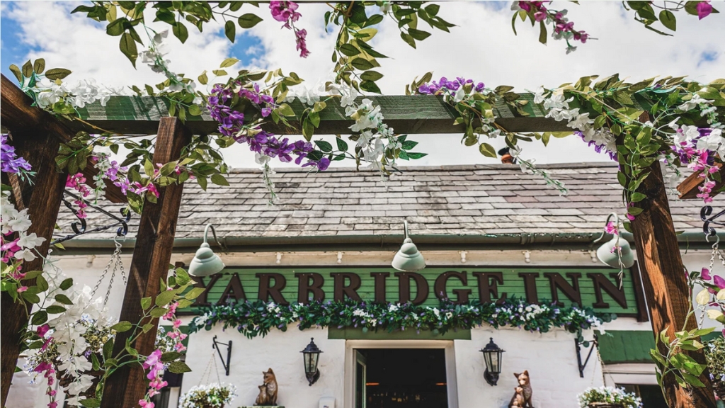 Decorative floral archway framing the entrance of the Yarbridge Inn in Brading, with colourful hanging flowers and a rustic exterior beneath a bright sky.