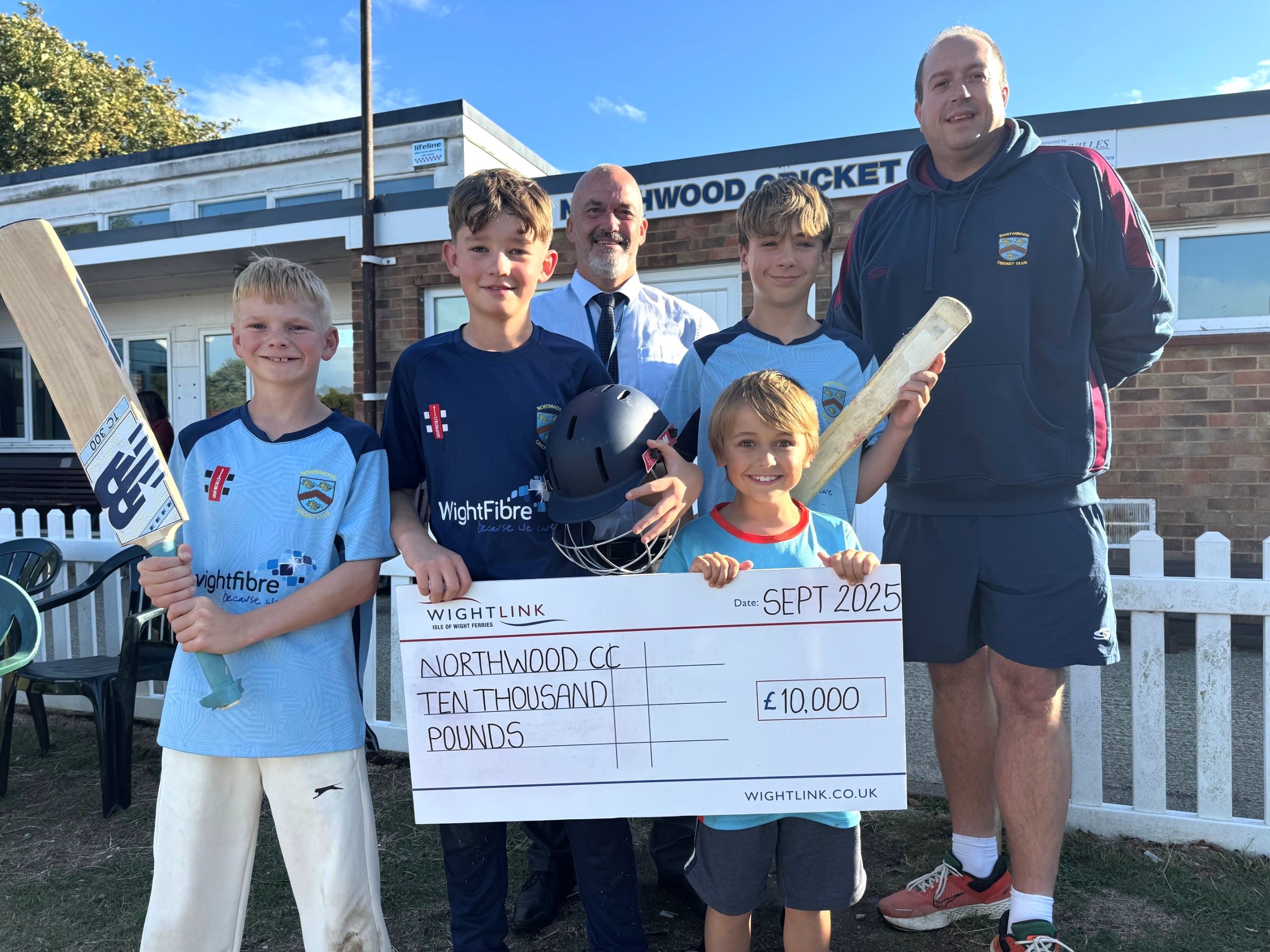 A group of young Northwood Cricket Club players and two adults stand smiling outside the clubhouse, holding cricket bats and a large presentation cheque from Wightlink for £10,000 dated September 2025.