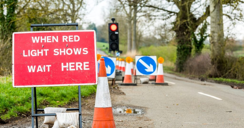 Temporary traffic lights and roadworks on a rural road, with a red sign reading “When red light shows wait here” beside cones and barriers controlling traffic flow.