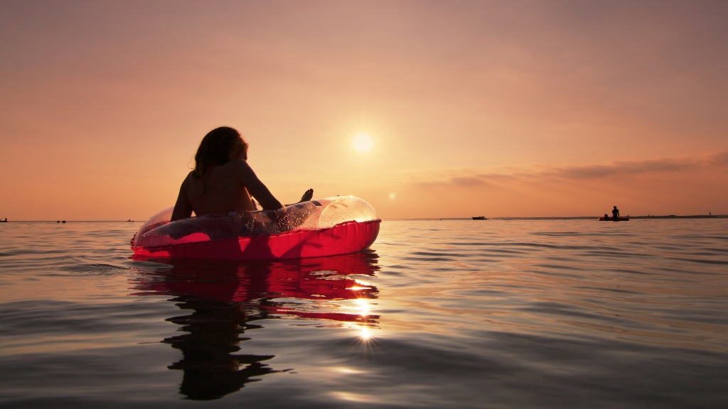. A person relaxes on an inflatable ring in calm sea water at sunset, with the sun low on the horizon and gentle reflections on the surface.
