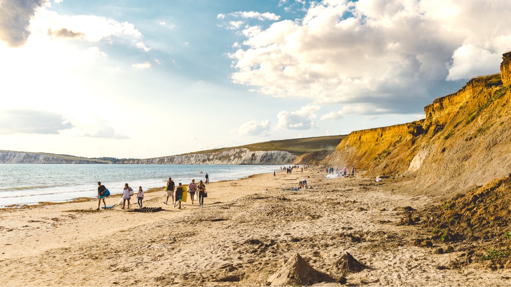People walk and relax along a wide sandy beach backed by dramatic cliffs, under a bright sky with scattered clouds.