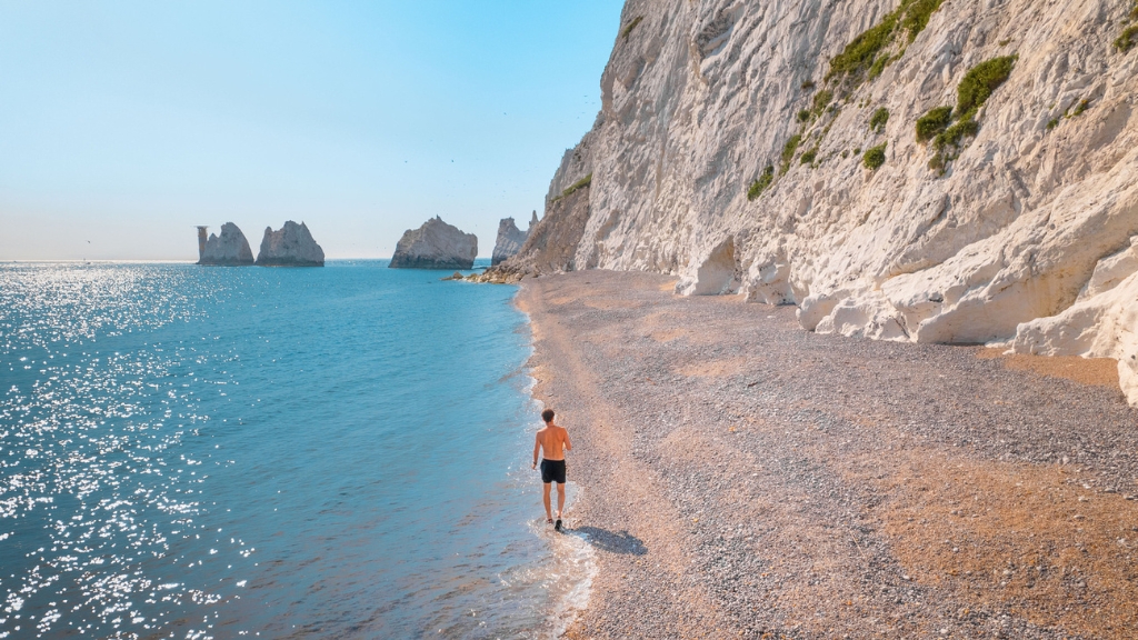 A person walks along a pebbled shoreline beneath towering white chalk cliffs, with the Needles rock formations visible offshore.