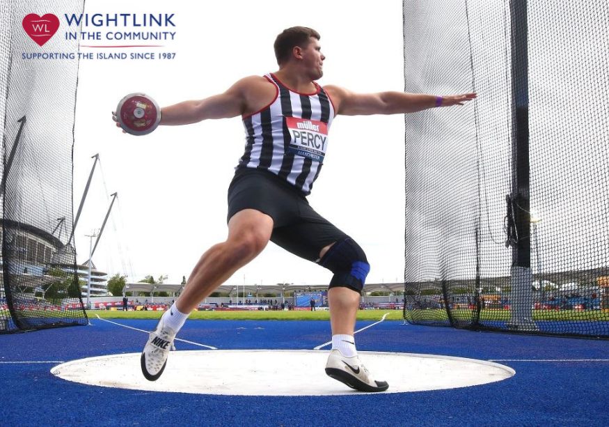 A male discus thrower in a black-and-white striped singlet labeled “PERCY” spins inside a throwing cage on a blue track, holding a discus with one arm extended, wearing a knee brace, with a stadium and Wightlink logo in the background.