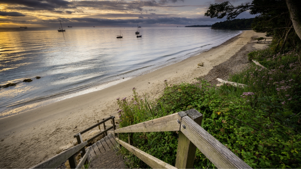 Wooden steps lead down to a quiet sandy bay at dusk, with anchored boats on calm water and trees framing the shoreline.