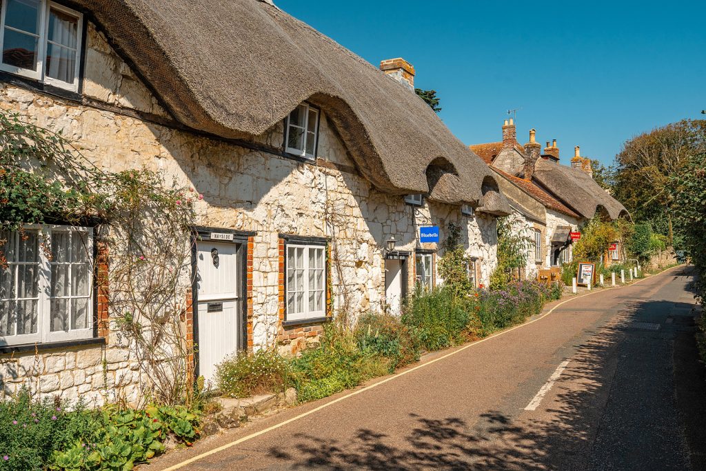 Row of traditional thatched stone cottages along a quiet village road on the Isle of Wight, with whitewashed walls, cottage gardens and blue sky.