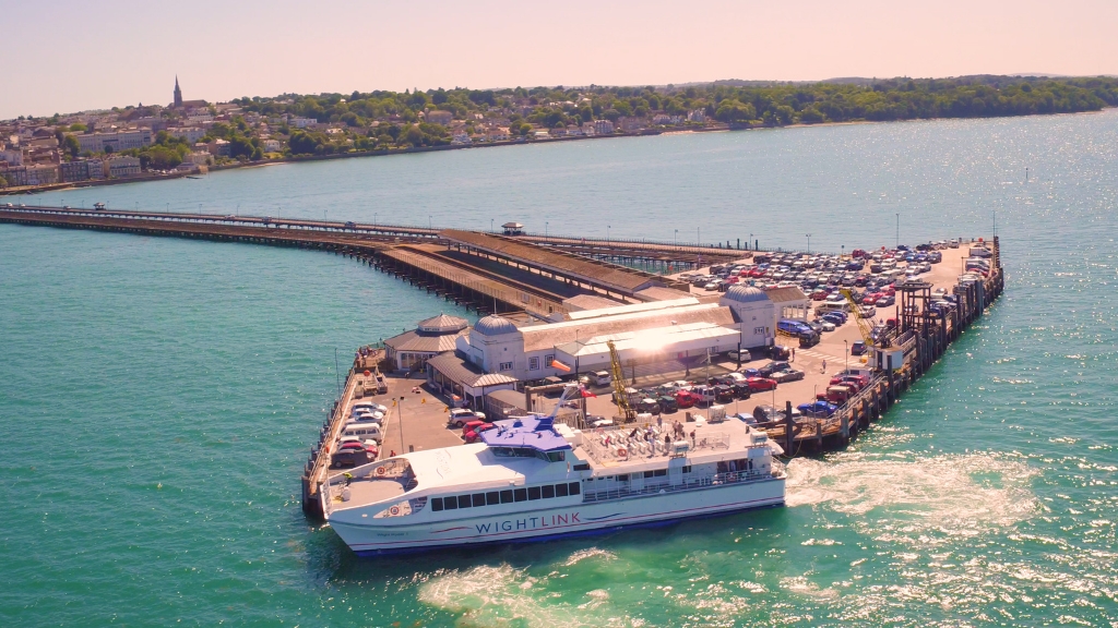 An aerial view of Ryde Pier and ferry terminal, with a Wightlink fast passenger ferry arriving alongside parked cars and turquoise water.
