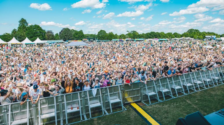 A large festival crowd stands packed together behind a barrier, cheering and raising their hands during a daytime outdoor concert.