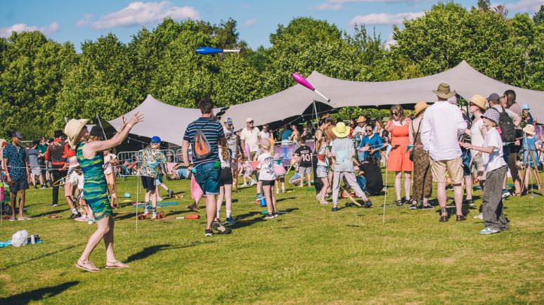 Families and festivalgoers enjoy activities on a grassy field, with tents in the background and people juggling and playing games.