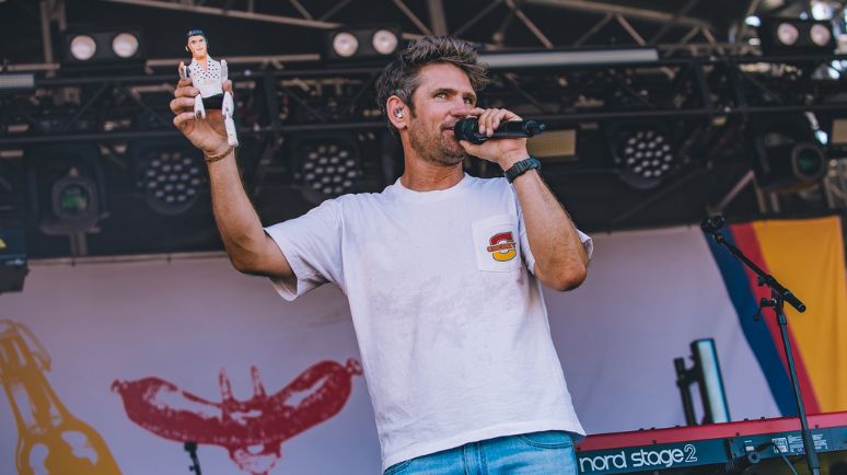 A singer addresses the crowd from the main stage, holding a microphone and a small figure, with stage lights and instruments behind him.