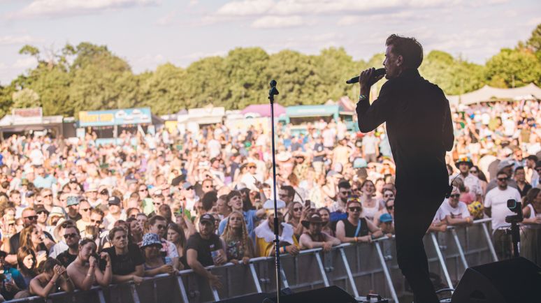 A performer sings on stage facing a large audience at an outdoor festival, with the crowd watching closely from behind the barrier.