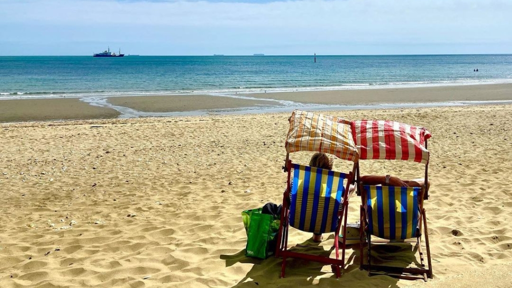 Two striped deckchairs sit on a sandy beach facing the sea, with calm blue water and ships visible on the horizon.