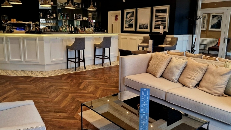 A stylish hotel bar and lounge area with a white bar counter, high stools, soft seating and framed artwork on dark walls.