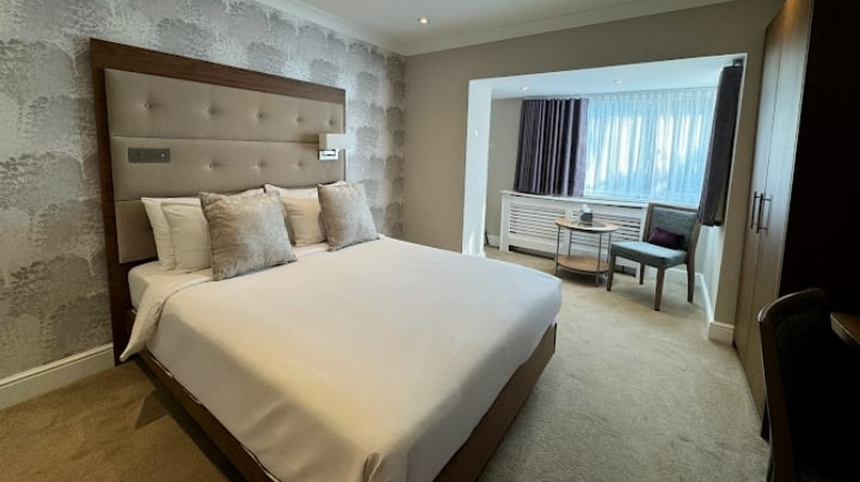 A modern hotel bedroom with a double bed, padded headboard, neutral décor and a seating area by the window.