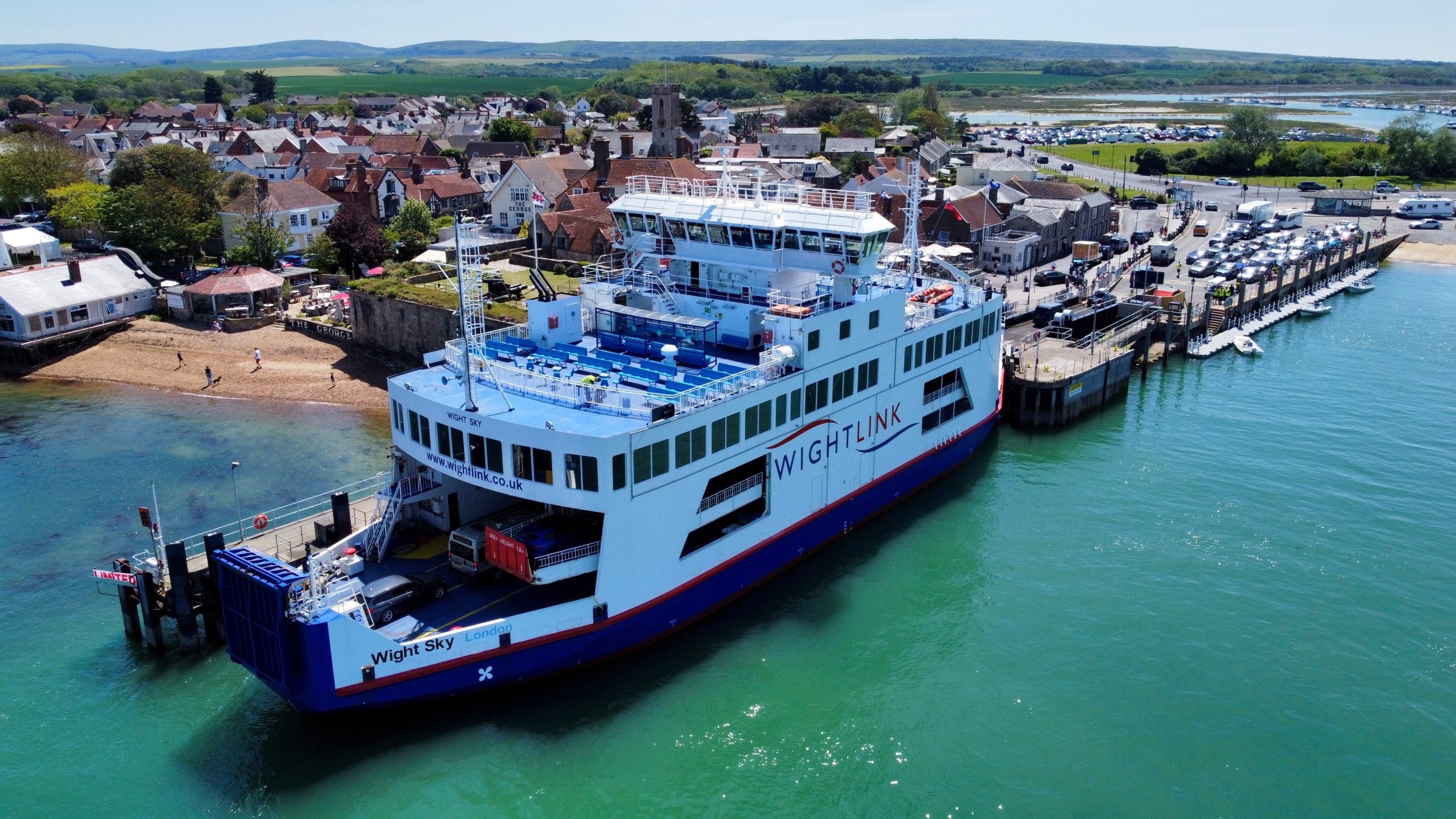 Aerial view of a Wightlink car ferry docked at Yarmouth on the Isle of Wight, with cars boarding, turquoise water, and a coastal town and countryside in the background.