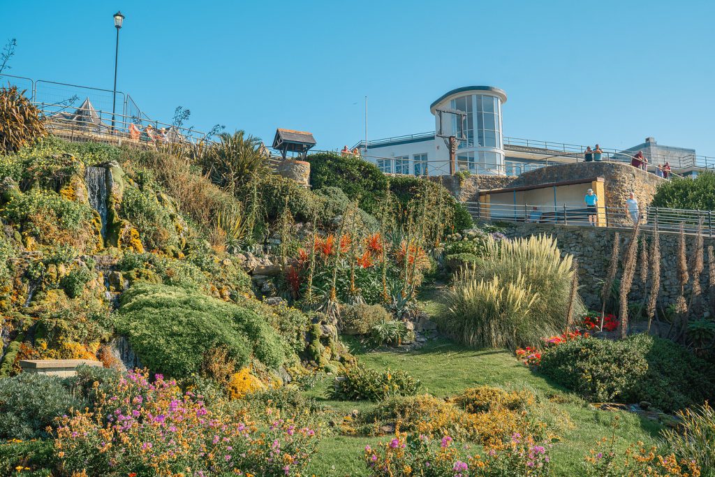 Cascade Gardens in Ventnor on the Isle of Wight, with colourful flowers, lush greenery and stone terraces leading up to a coastal promenade and art deco style building above.