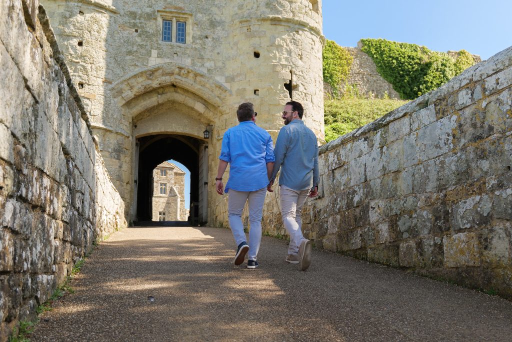 Two people walk hand in hand along the stone pathway into Carisbrooke Castle, framed by historic walls and a medieval archway on the Isle of Wight.