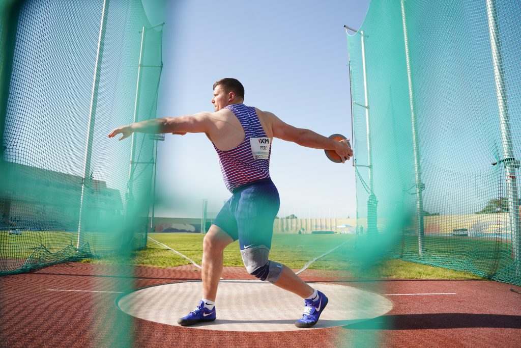 A male discus thrower in a black-and-white striped singlet labeled “PERCY” spins inside a throwing cage on a blue track, holding a discus with one arm extended, wearing a knee brace, with a stadium and Wightlink logo in the background.