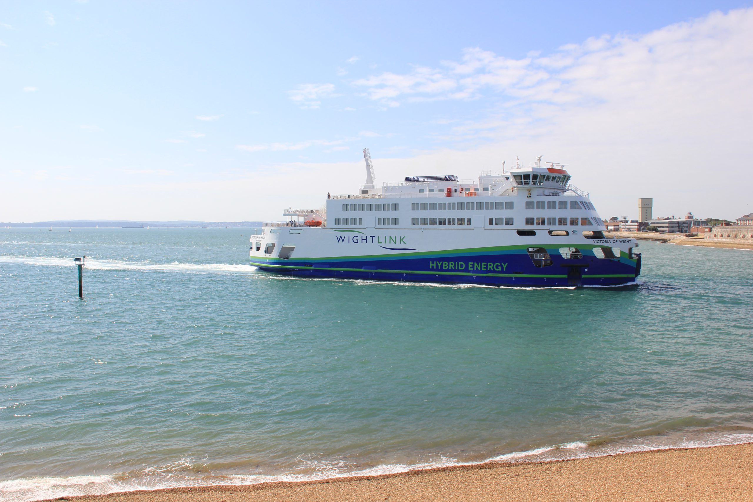 A large white and blue hybrid ferry, labeled "WIGHTLINK" and "HYBRID ENERGY," sails through the calm blue water near a shoreline. In the background, a part of a coastal town is visible, with a water tower and buildings along the beach. The sky is bright and partly cloudy.