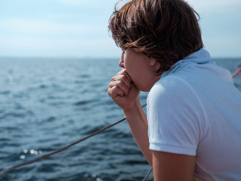 A young man wearing a white t-shirt is looking out to sea from a boat, he has his hand in front of his mouth and looks seasick