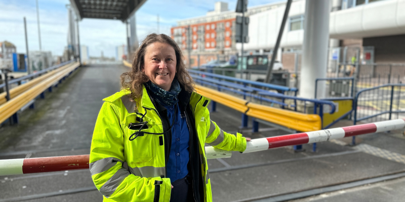 A woman wearing a blue shirt and a neon yellow high visibility coat standing against a barrier to a ferry entrance